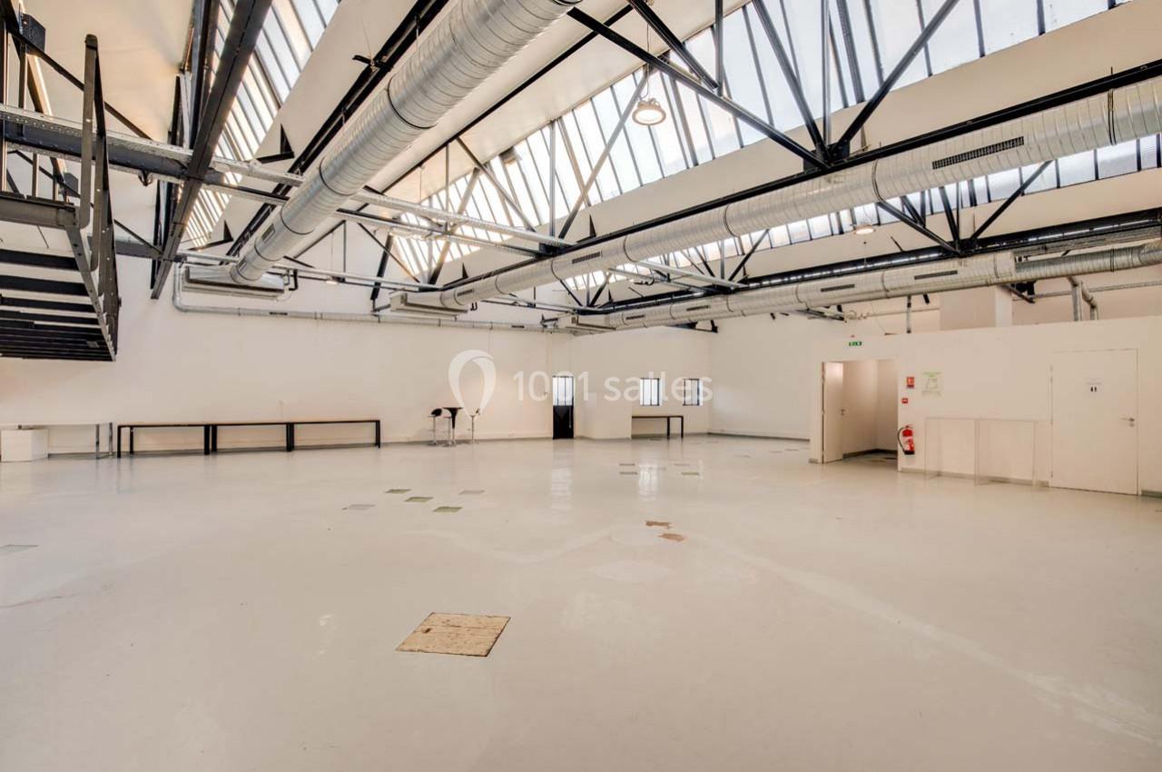 Intérieur d'un grand espace industriel lumineux avec toit en verrière, poutres métalliques et sol clair.