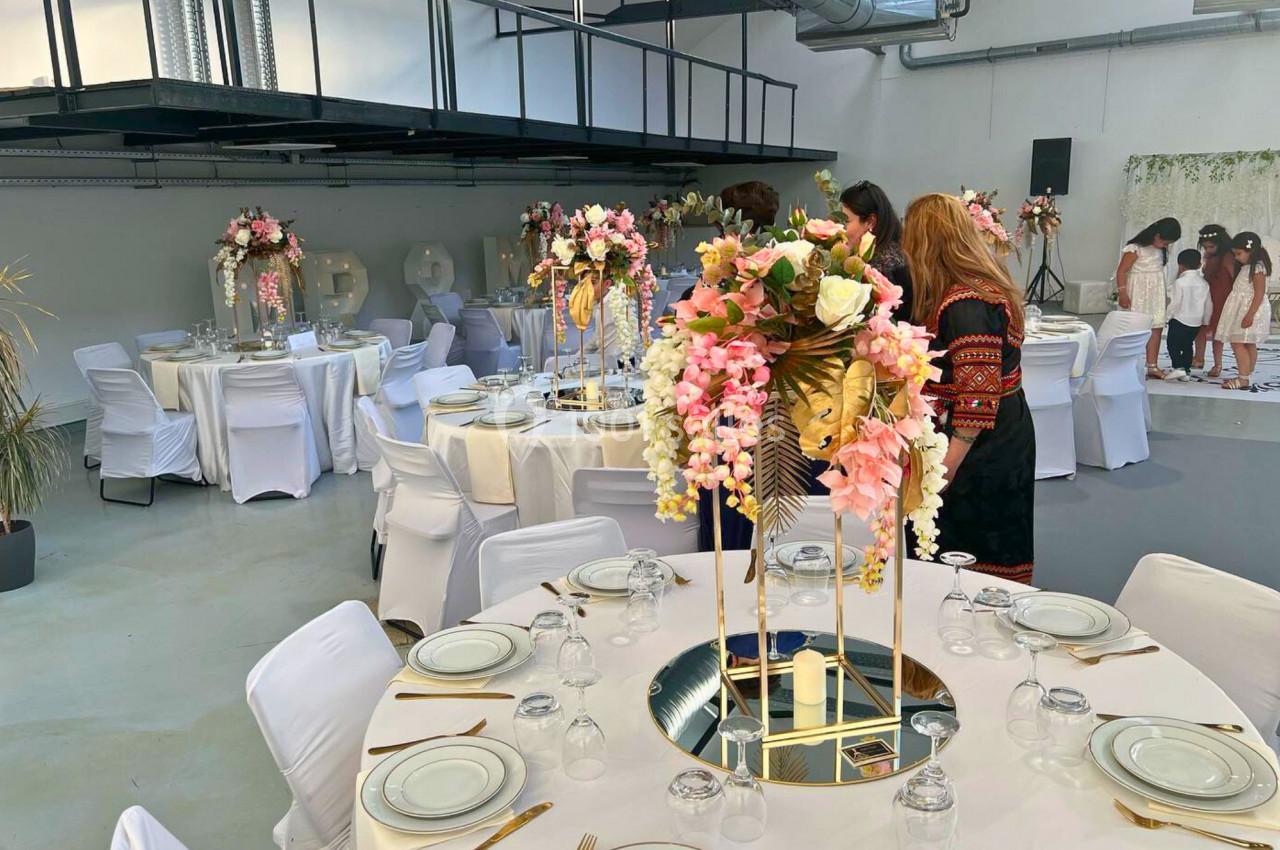 Salle de réception décorée avec des tables rondes, nappes blanches, centres de table fleuris et chaises houssées.