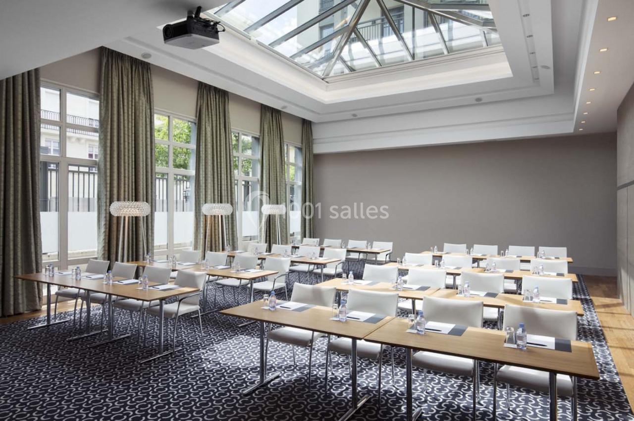 Salle de conférence lumineuse avec des tables alignées, des chaises blanches et un plafond vitré.