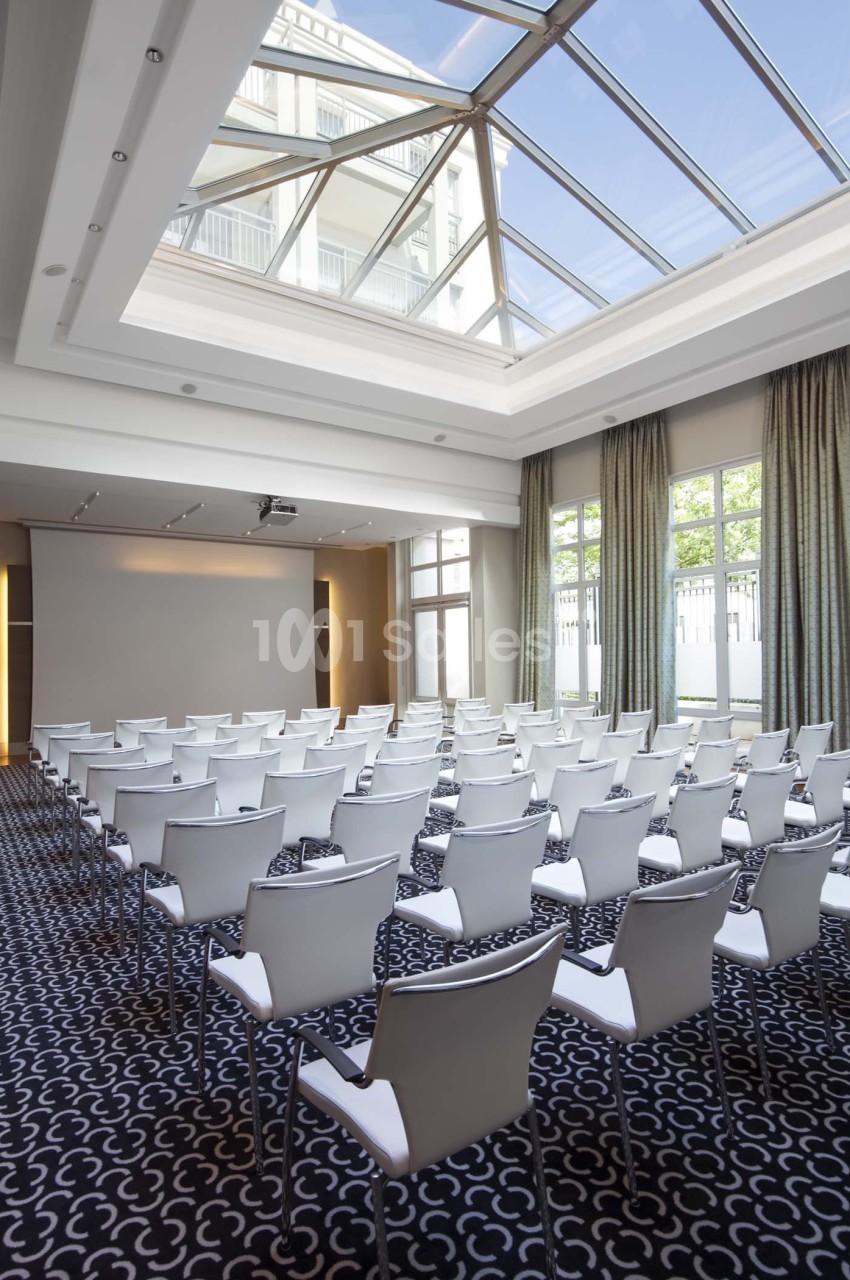 Salle de conférence lumineuse avec des chaises blanches alignées, un écran de projection et une verrière au plafond.