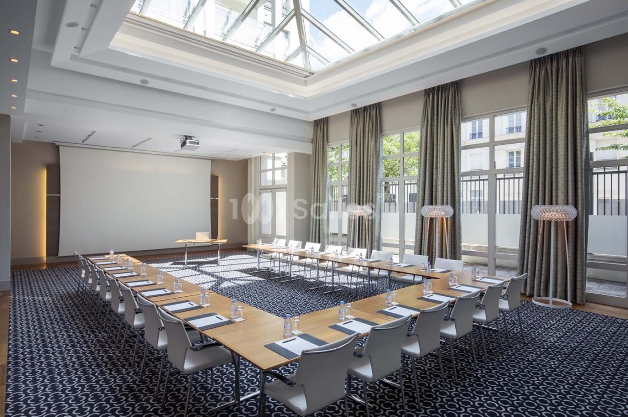 Salle de réunion lumineuse avec grandes baies vitrées, tables disposées en U et chaises blanches.