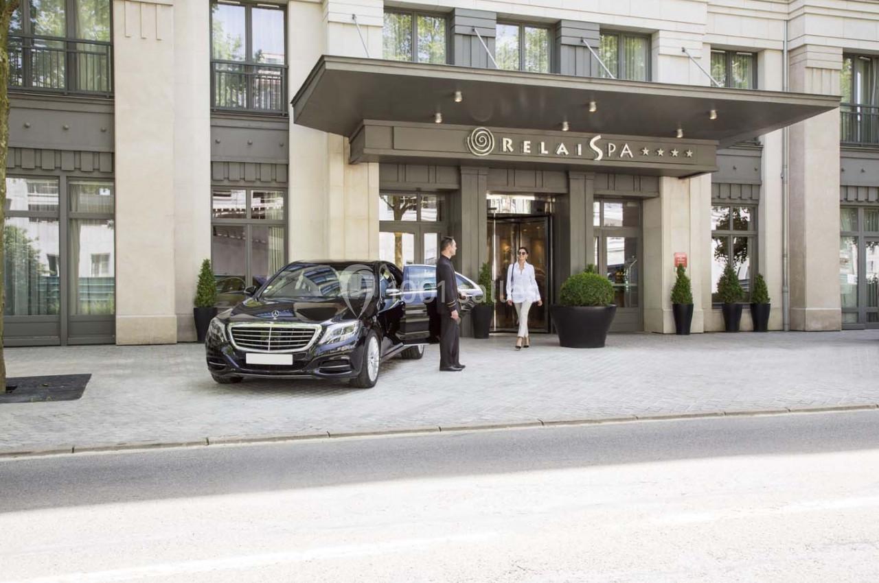 Entrée d'un hôtel avec une voiture noire garée devant, un chauffeur ouvrant la porte pour une cliente.