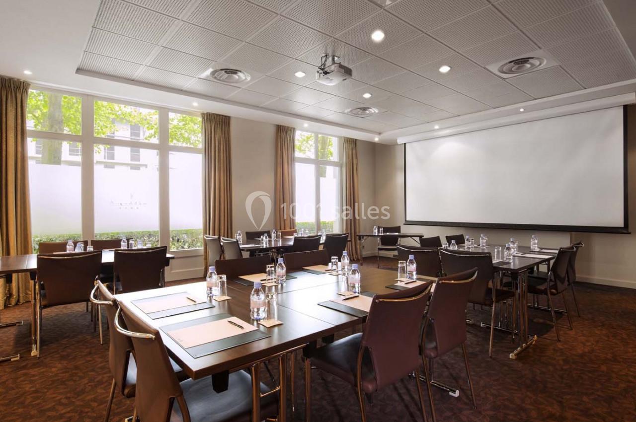 Salle de réunion lumineuse avec tables disposées en rangées, chaises, bouteilles d'eau et écran de projection.