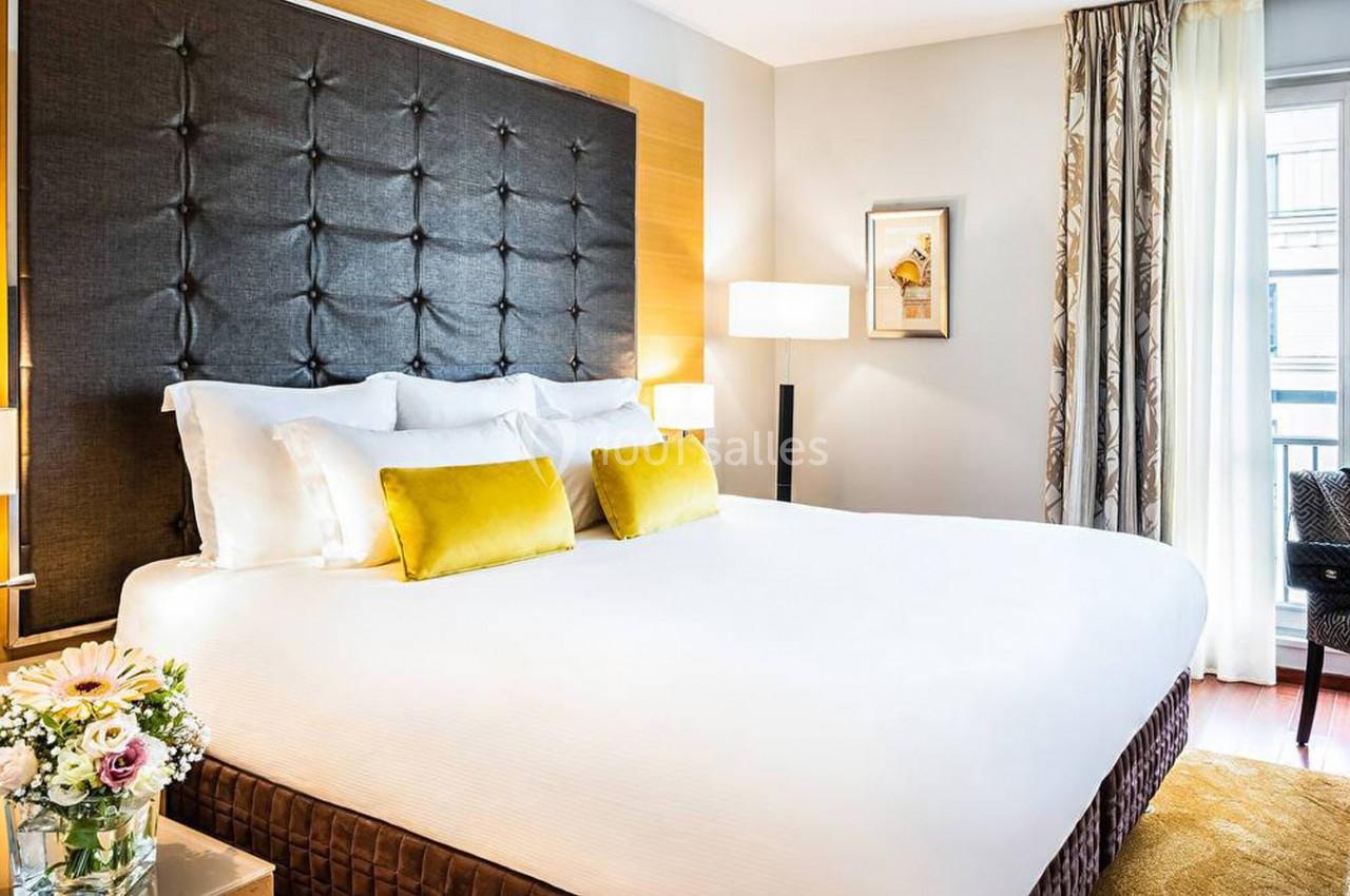 Chambre lumineuse avec un grand lit, tête de lit capitonnée, coussins jaunes et vue sur une fenêtre avec rideaux.