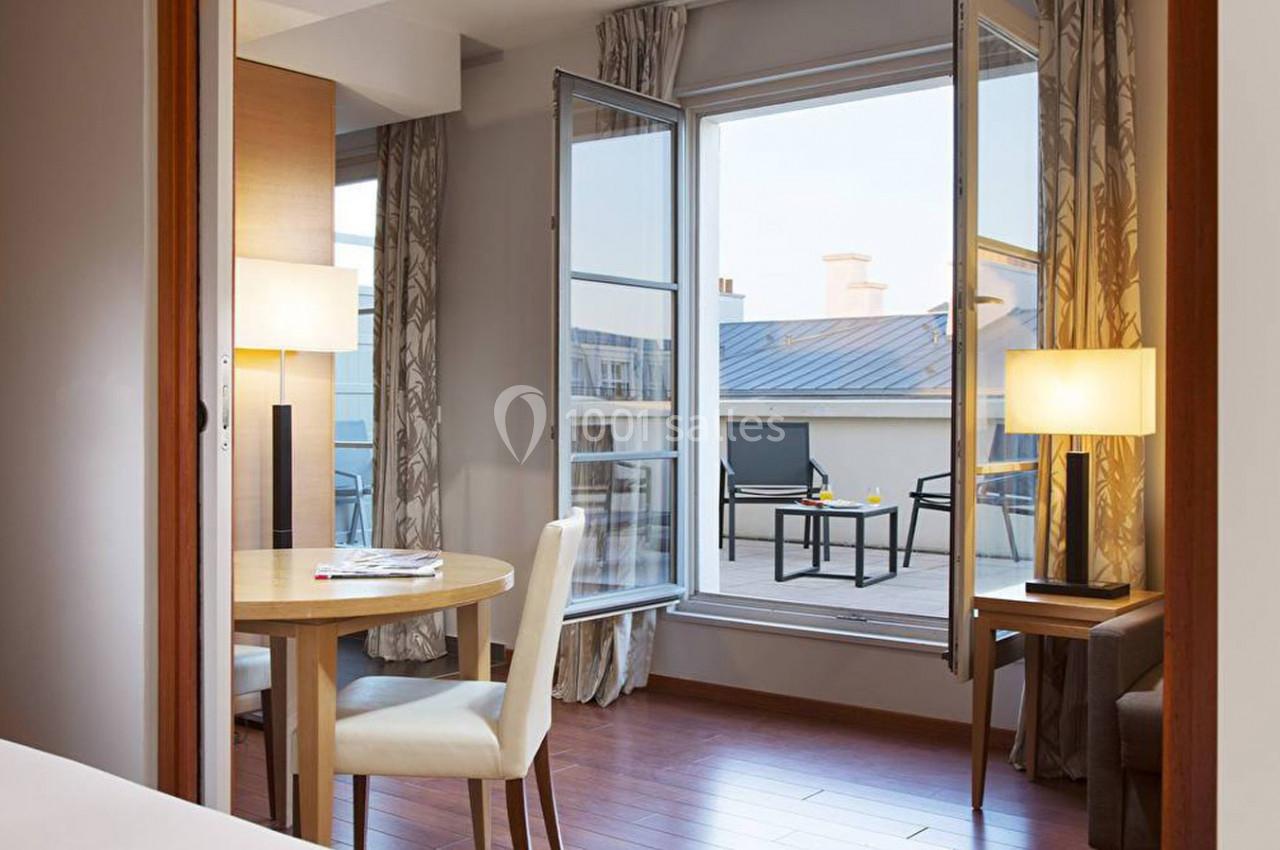 Vue d'une chambre lumineuse avec table, chaises, lampes et accès à une terrasse extérieure avec mobilier.