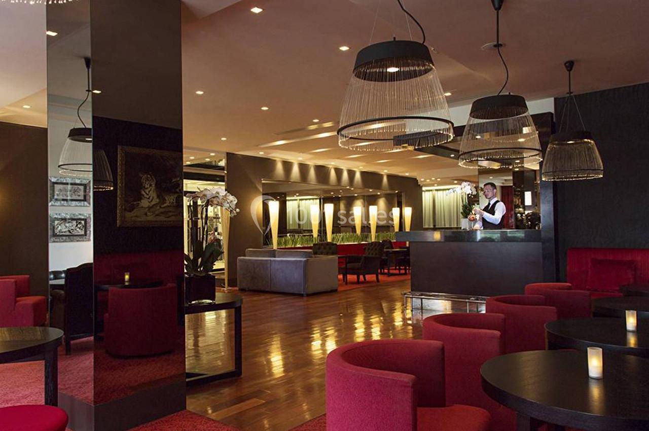 Salle de restaurant élégante avec tables noires, fauteuils rouges, éclairage tamisé et un serveur au comptoir.