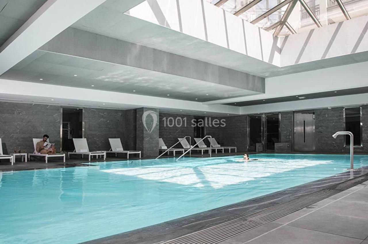 Piscine intérieure lumineuse entourée de chaises longues, avec un plafond en verre laissant entrer la lumière naturelle.