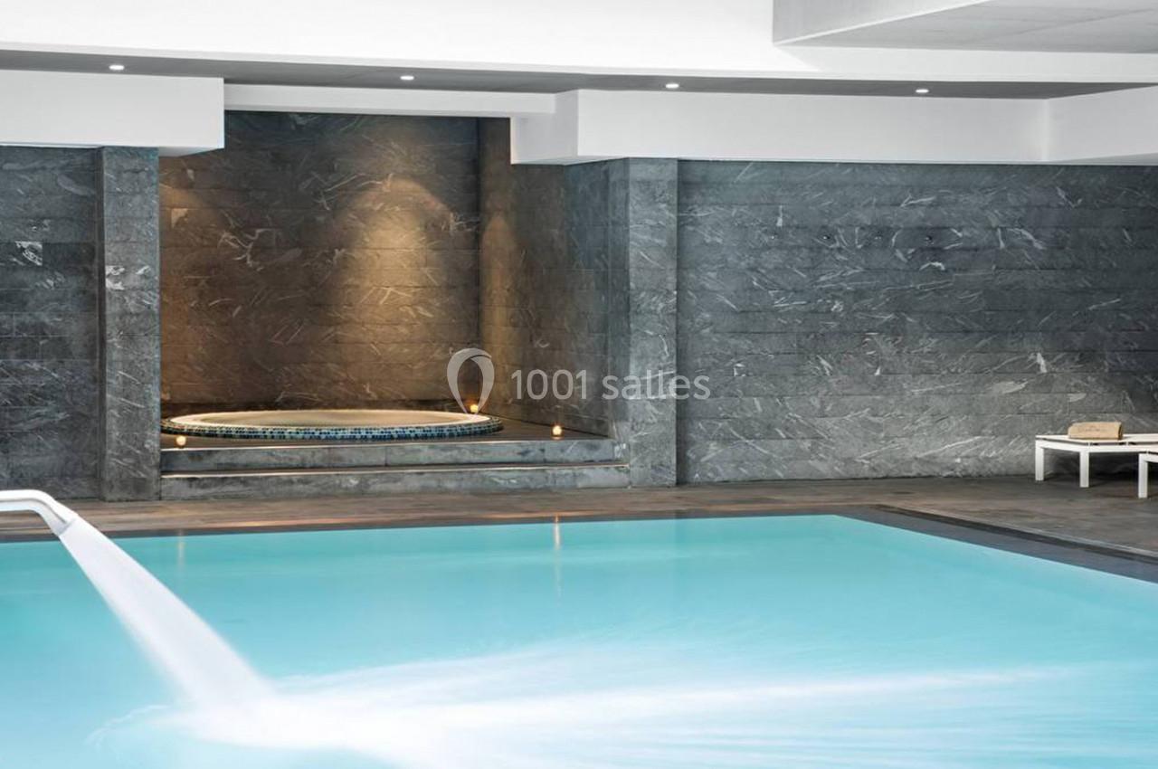 Piscine intérieure avec cascade d'eau, jacuzzi encastré dans un mur en pierre et transat blanc sur le côté.