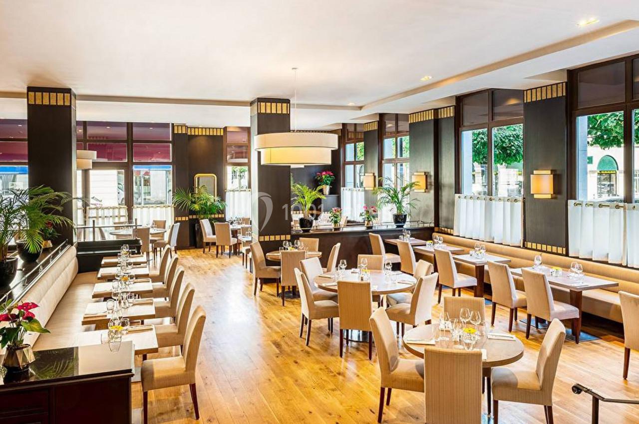 Salle de restaurant lumineuse avec tables dressées, chaises en bois clair, grandes fenêtres et décorations végétales.
