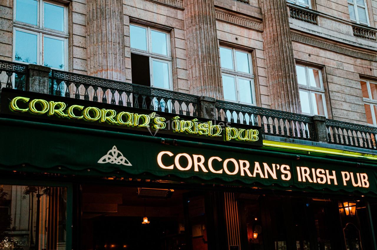 Location salle Paris 2 (Paris) - Corcoran's Grands Boulevards #12 Façade d'un pub irlandais avec enseignes lumineuses vertes et jaunes, devant un bâtiment en pierre.