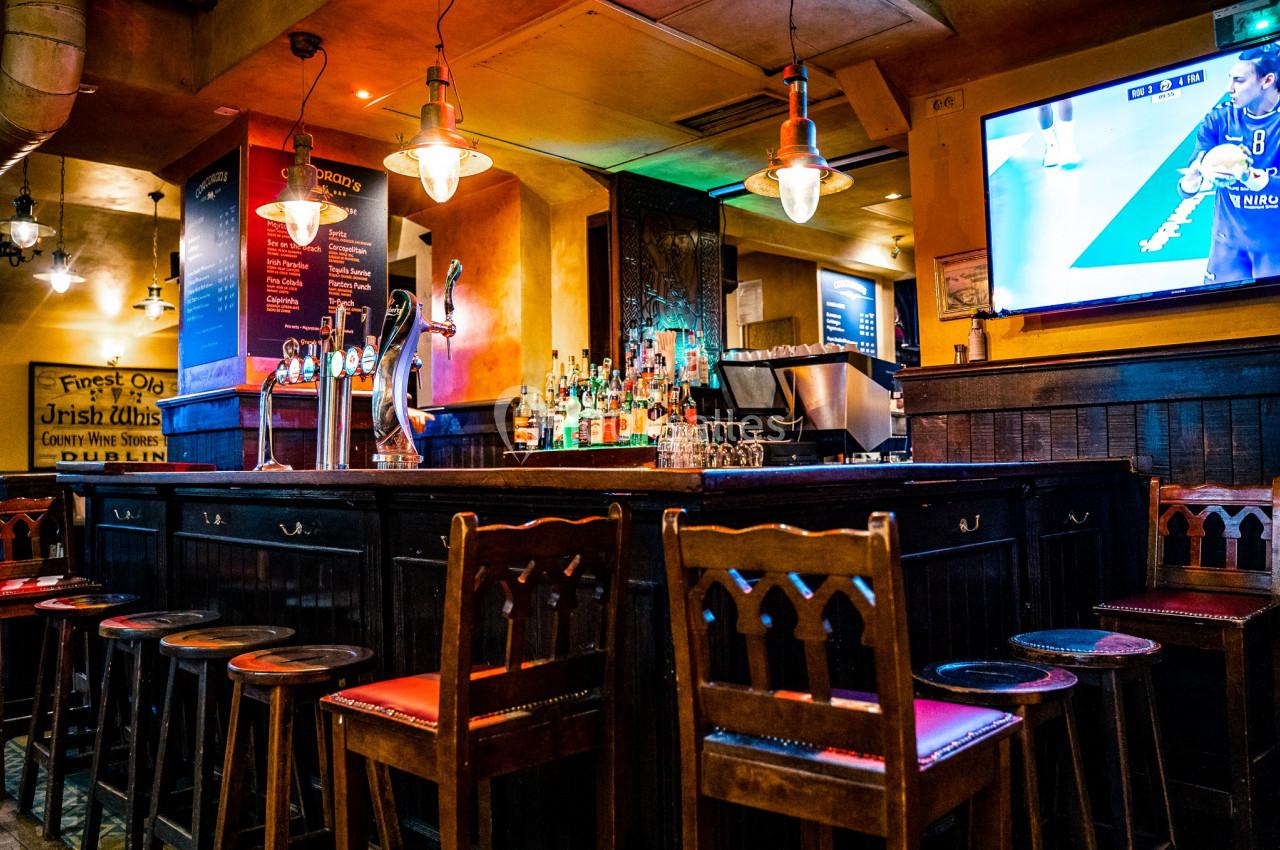 Location salle Paris 2 (Paris) - Corcoran's Grands Boulevards #8 Intérieur d'un pub avec un bar en bois, des tabourets, des lumières suspendues et un écran diffusant un match.