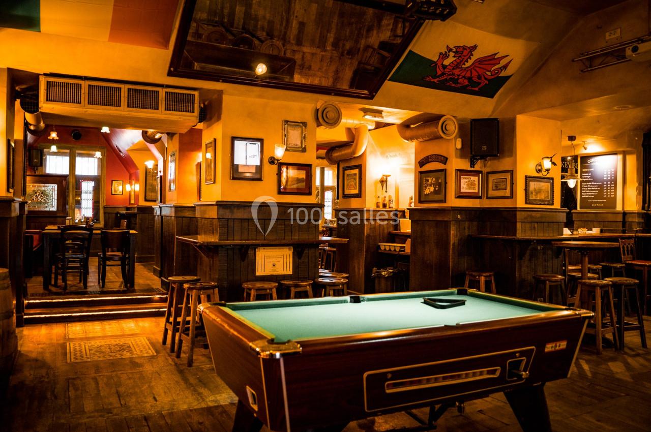 Salle de bar chaleureuse avec tables hautes, tabourets en bois et une table de billard au premier plan.