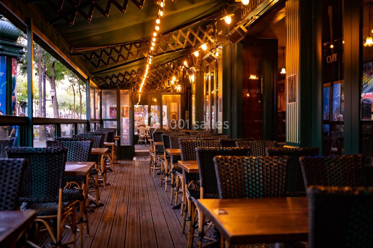 Terrasse de café vide avec des tables et chaises en osier, éclairée par des guirlandes lumineuses en soirée.