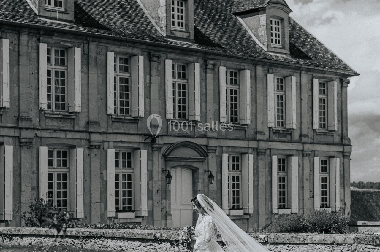 Location salle Wy-dit-Joli-Village (Val-d'Oise) - Château d'Hazeville #3 Une mariée en robe blanche marche sur une pelouse devant un grand bâtiment ancien en pierre.