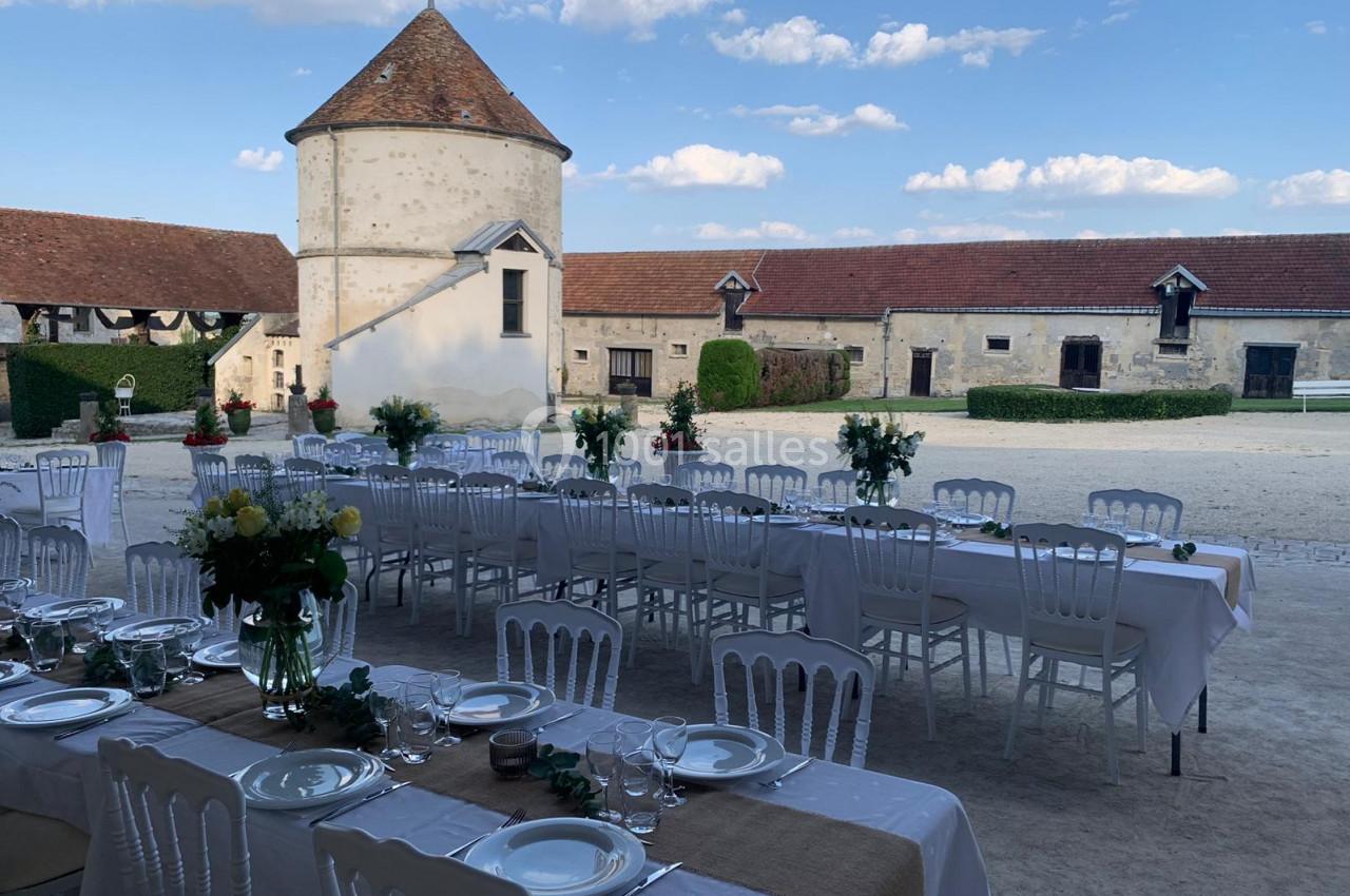 Location salle Wy-dit-Joli-Village (Val-d'Oise) - Château d'Hazeville #19 Tables dressées avec nappes blanches et décorations florales dans une cour de ferme avec bâtiments en pierre et ciel dégagé.