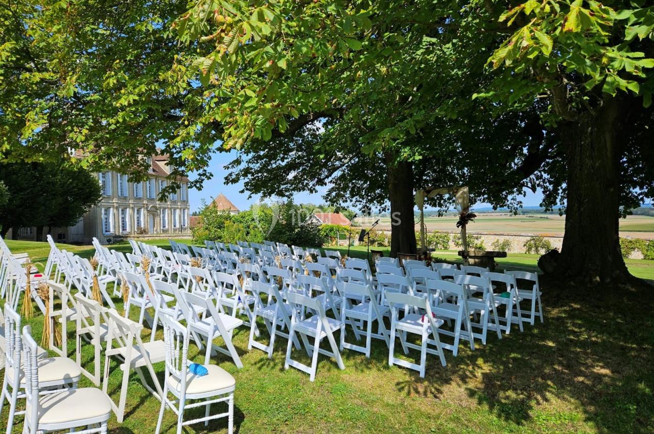 Location salle Wy-dit-Joli-Village (Val-d'Oise) - Château d'Hazeville #21 Chaises blanches alignées en extérieur sous des arbres, préparées pour une cérémonie avec vue sur un paysage champêtre.