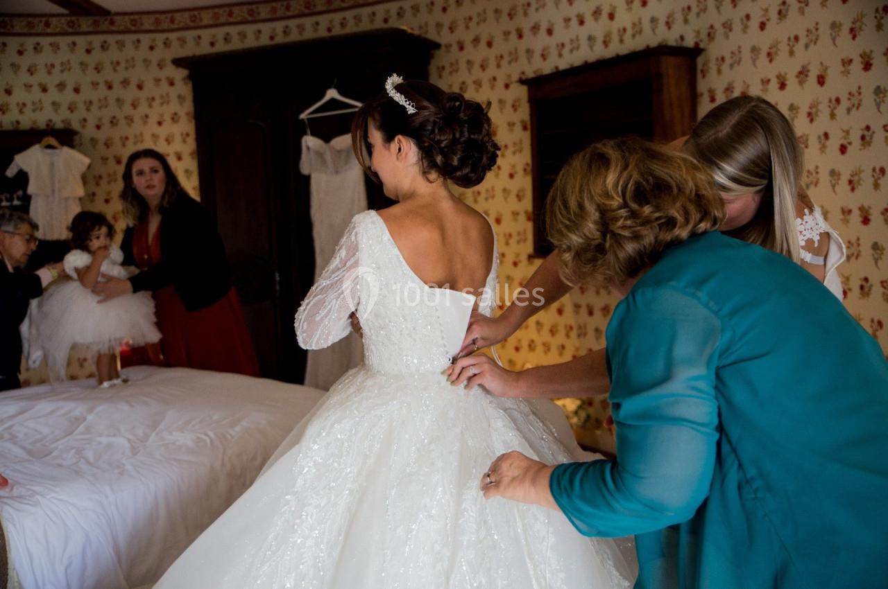 Location salle Wy-dit-Joli-Village (Val-d'Oise) - Château d'Hazeville #22 Une mariée en robe blanche est aidée par deux femmes pour ajuster sa tenue dans une chambre décorée de papier peint fleuri.