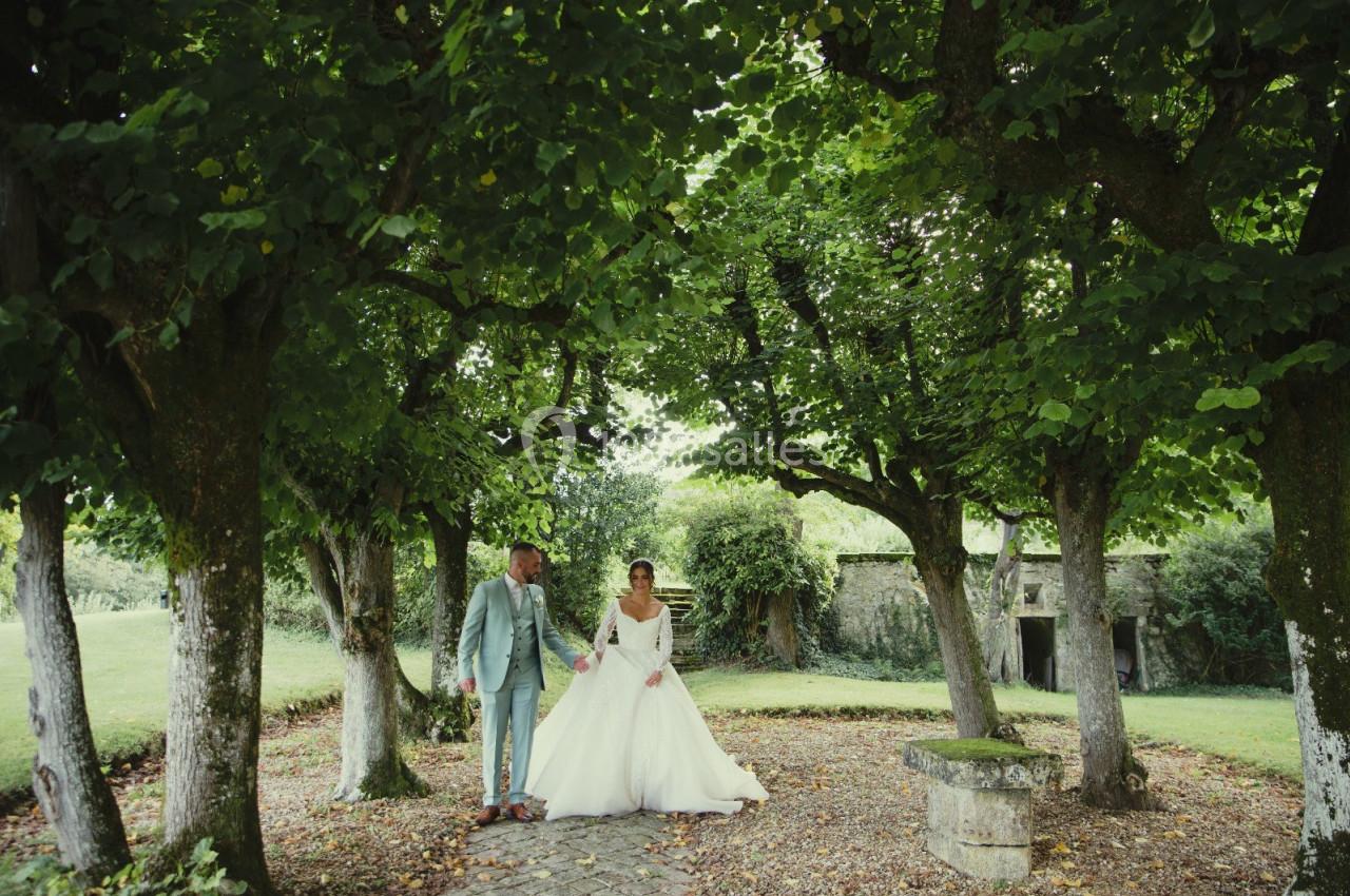 Location salle Wy-dit-Joli-Village (Val-d'Oise) - Château d'Hazeville #26 Un couple en tenue de mariage pose sous des arbres dans un jardin verdoyant avec un banc en pierre à proximité.