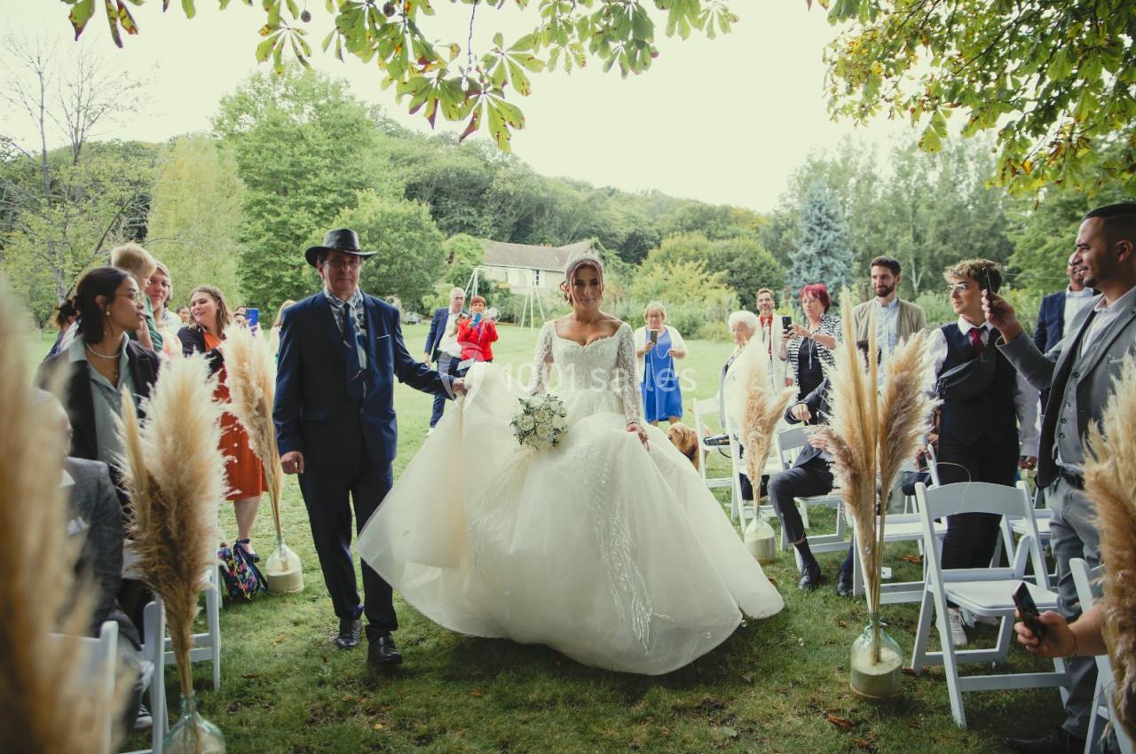 Location salle Wy-dit-Joli-Village (Val-d'Oise) - Château d'Hazeville #30 Une mariée en robe blanche avance dans une allée extérieure bordée d'invités assis sous des arbres.