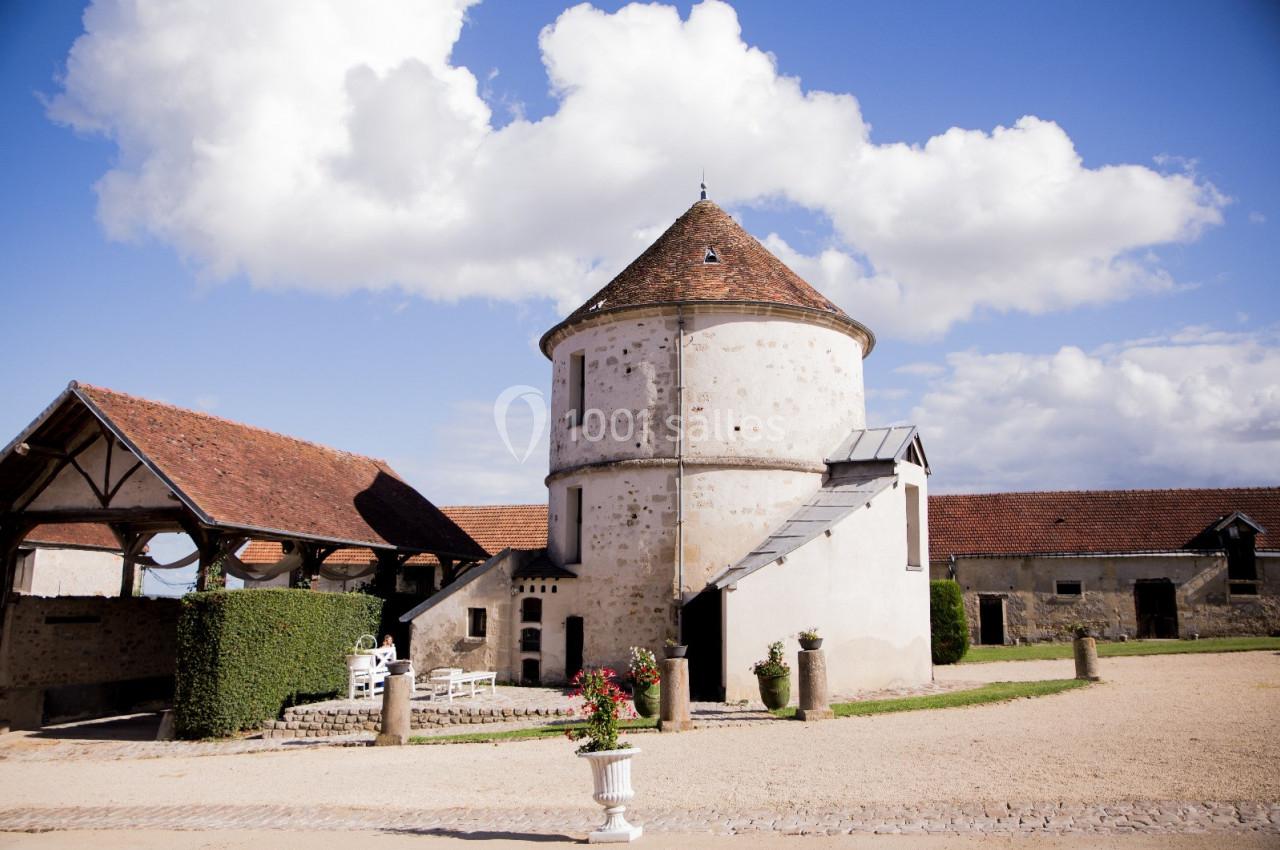 Location salle Wy-dit-Joli-Village (Val-d'Oise) - Château d'Hazeville #31 Bâtiment en pierre avec toit en tuiles, entouré de dépendances et d'une cour gravillonnée sous un ciel bleu.