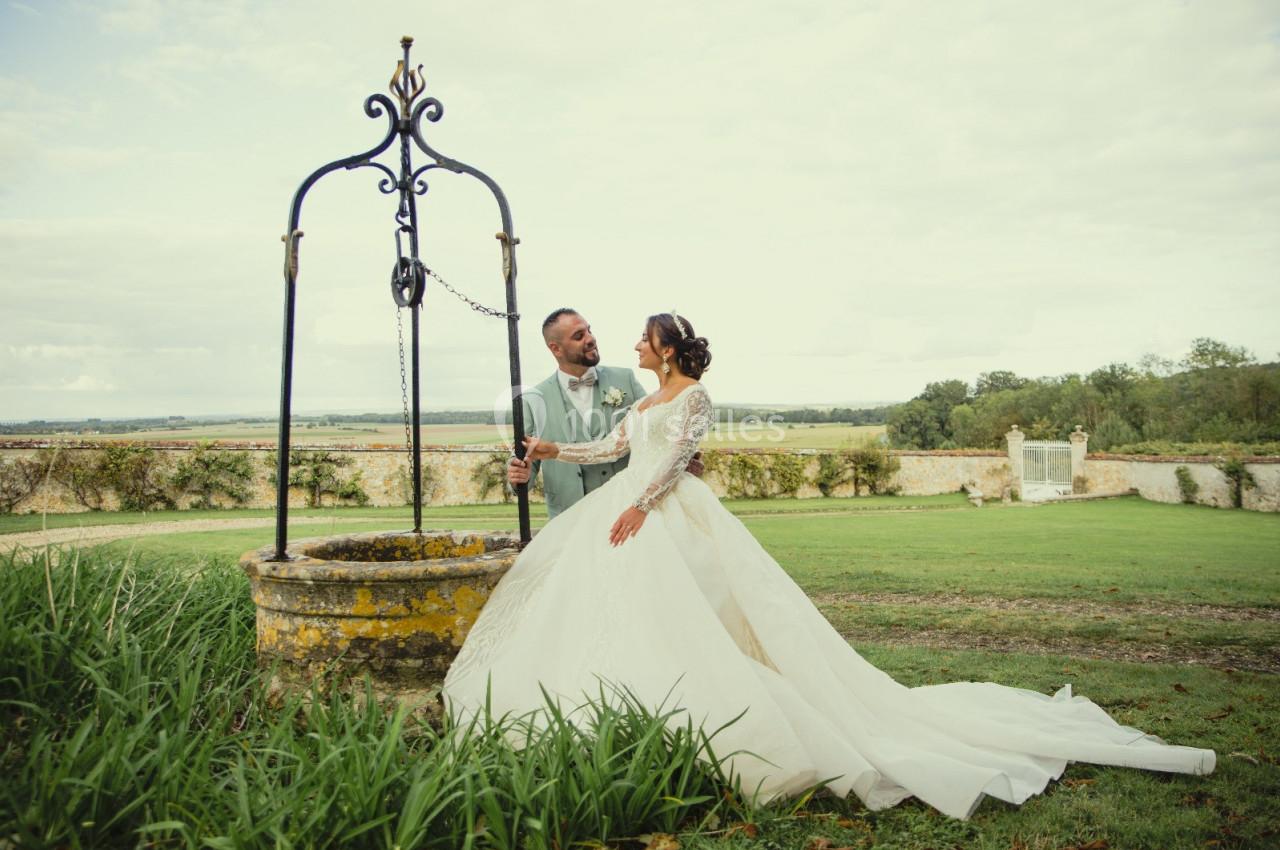 Location salle Wy-dit-Joli-Village (Val-d'Oise) - Château d'Hazeville #24 Un couple en tenue de mariage se tient près d'un puits ancien dans un jardin verdoyant.