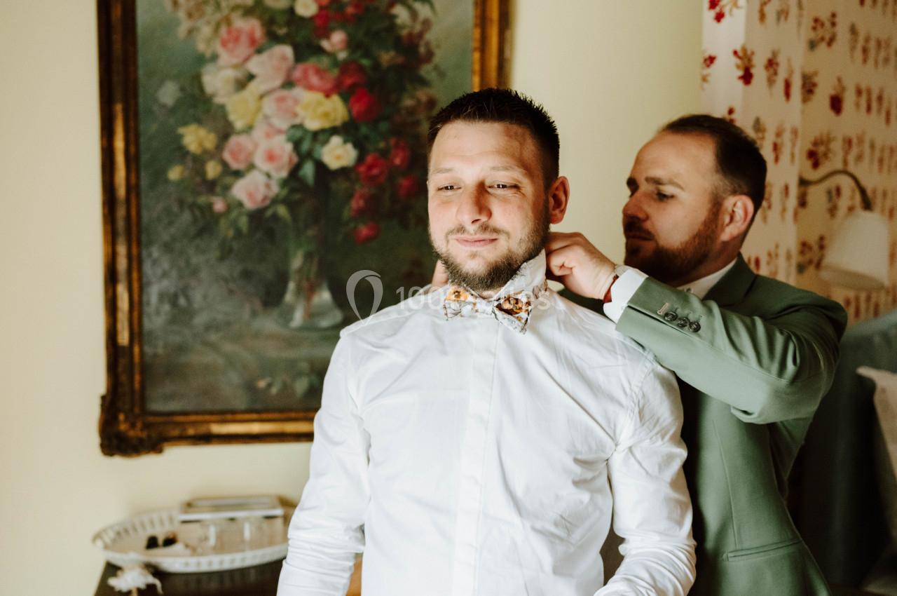 Location salle Wy-dit-Joli-Village (Val-d'Oise) - Château d'Hazeville #35 Un homme ajuste le nœud papillon d'un autre homme dans une pièce décorée avec un tableau floral et des motifs rouges.