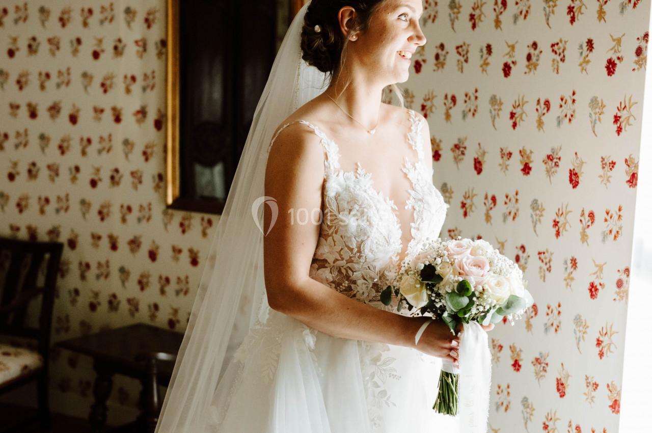 Location salle Wy-dit-Joli-Village (Val-d'Oise) - Château d'Hazeville #36 Une mariée en robe blanche tient un bouquet de fleurs, debout près d'une fenêtre dans une pièce au papier peint fleuri.