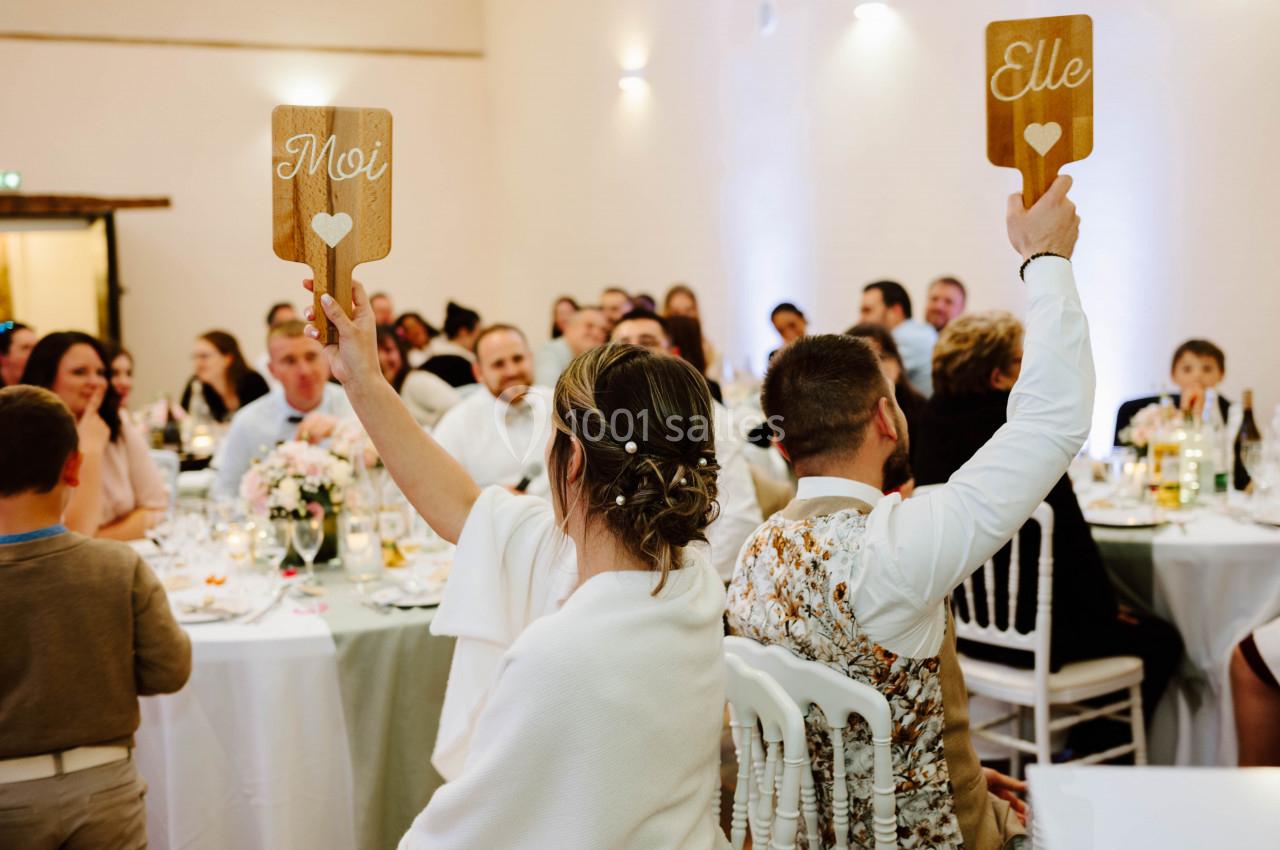 Location salle Wy-dit-Joli-Village (Val-d'Oise) - Château d'Hazeville #10 Un couple assis de dos lors d'un mariage lève des pancartes en bois indiquant ’Moi’ et ’Elle’ devant des invités.
