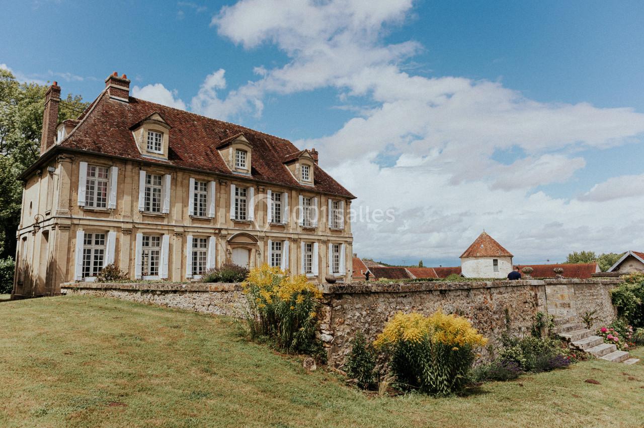 Chateau d'Hazeville Grande maison en pierre avec toit en tuiles, entourée d'un jardin fleuri et d'un mur en pierre sous un ciel partiellement…