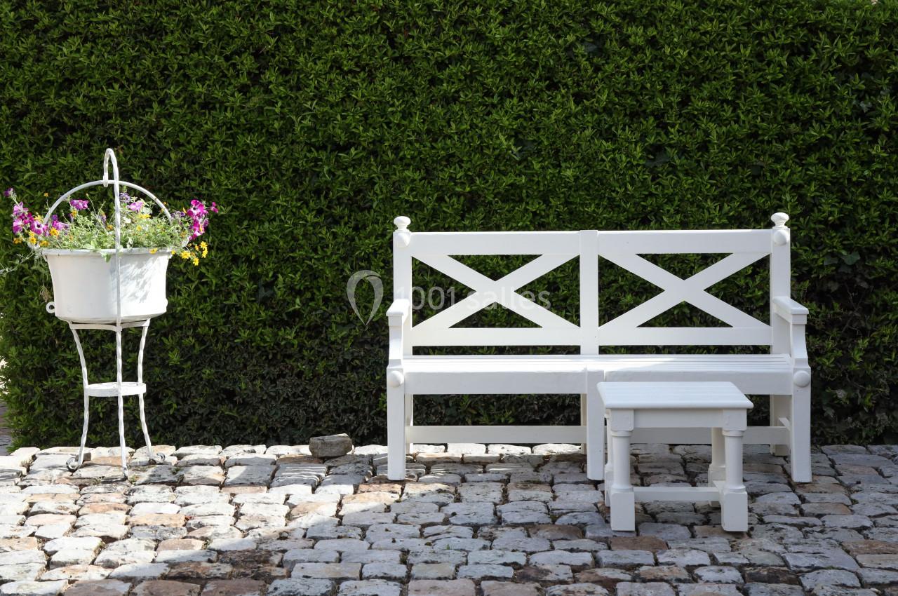 Des espaces cosy & intimistes Banc et table blanche en bois sur une terrasse pavée, avec un support de fleurs blanches et un mur de haie verte.