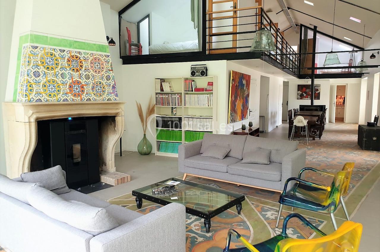 Grand loft famillial Salon spacieux avec cheminée ornée de carreaux colorés, canapés gris, chaises transparentes et mezzanine visible.