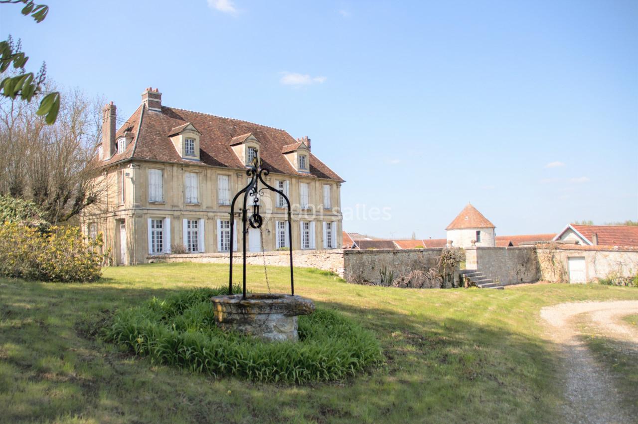 Parc bucolique et romantique Grande maison en pierre avec toit en tuiles, puits ancien au premier plan et jardin verdoyant sous un ciel dégagé.