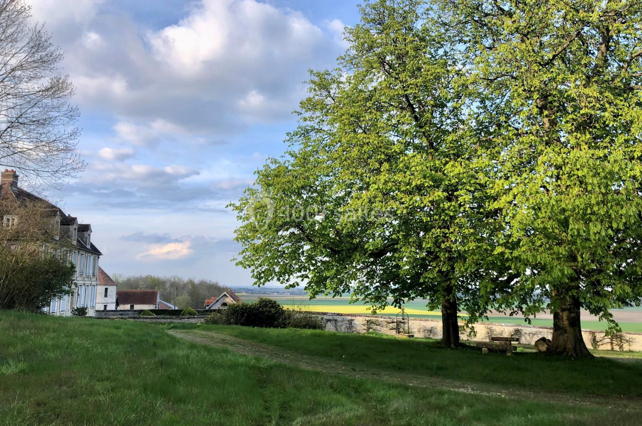 Parc du château - vue surplombant la plaine Maison ancienne près d'un grand arbre sur une pelouse verdoyante, avec un paysage champêtre en arrière-plan.