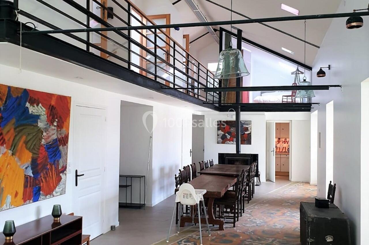 Séjour géant pour se retrouver en famille Intérieur lumineux avec mezzanine, grande table en bois, tapis coloré et œuvres d'art accrochées au mur.