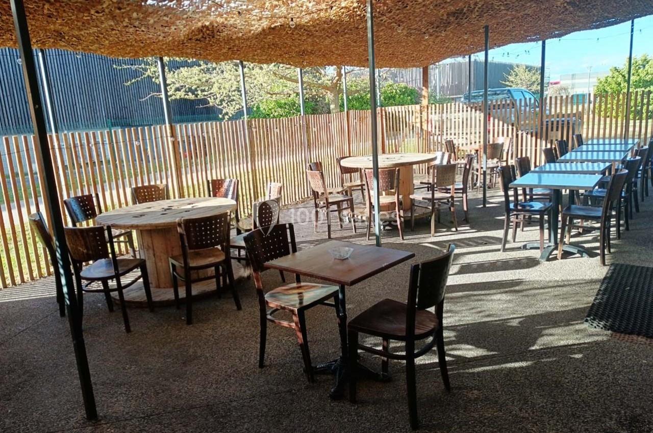 Terrasse extérieure ombragée avec des tables et chaises en bois, entourée d'une clôture en bois clair.