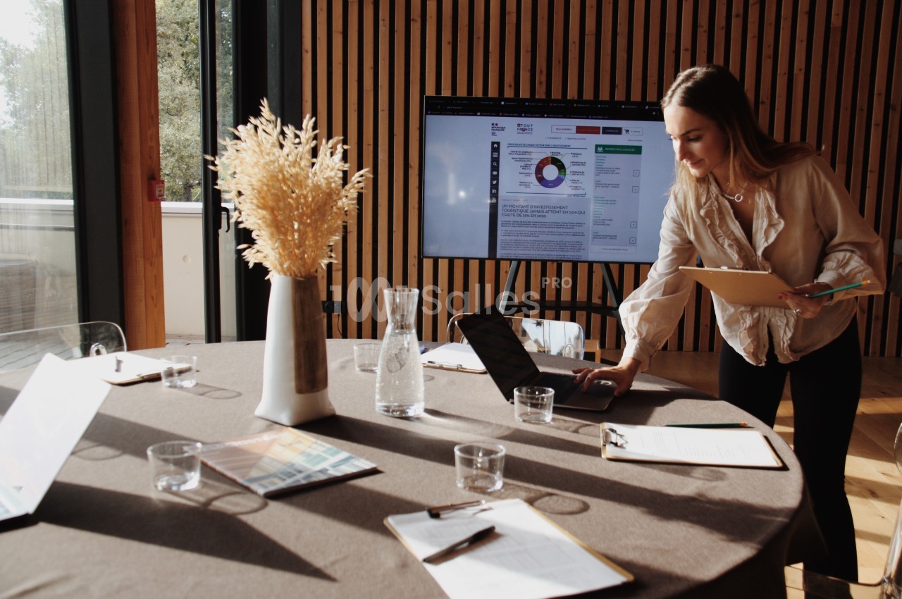 Une femme consulte un ordinateur portable sur une table de réunion avec des documents, un écran et un vase de fleurs séchées.