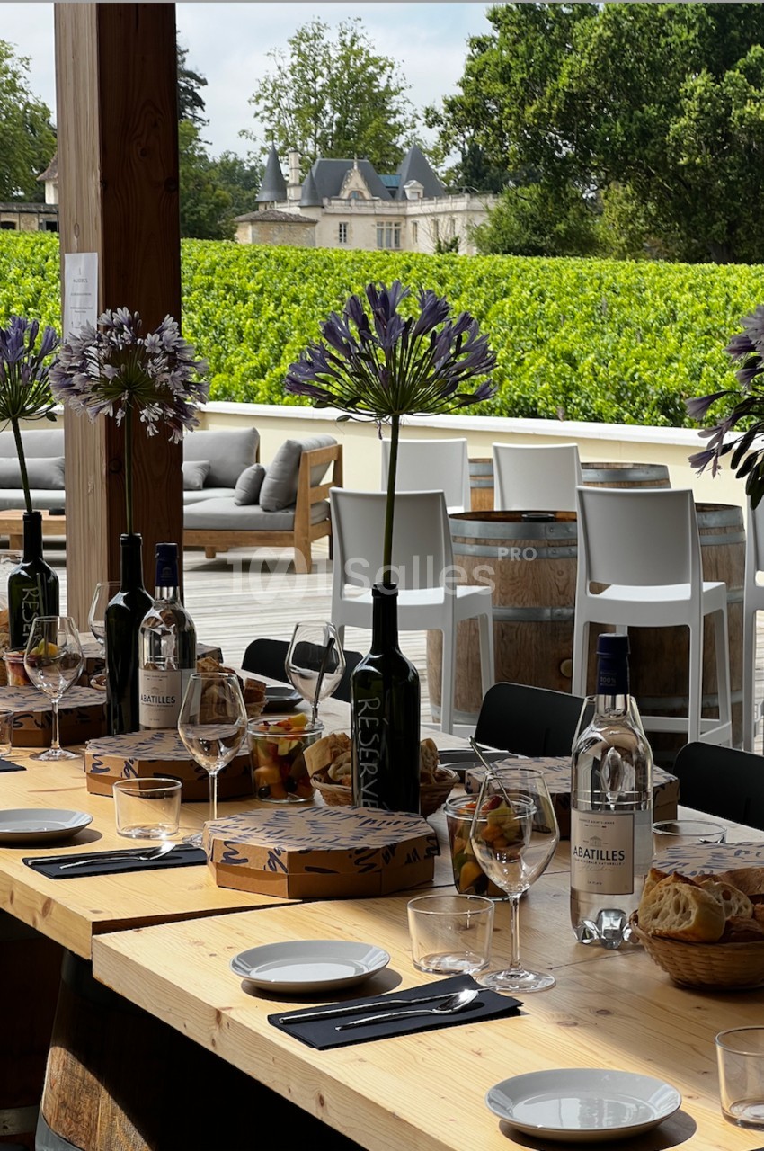 Table dressée en extérieur avec bouteilles de vin, verres, paniers de pain et fleurs, vue sur un vignoble et un château.
