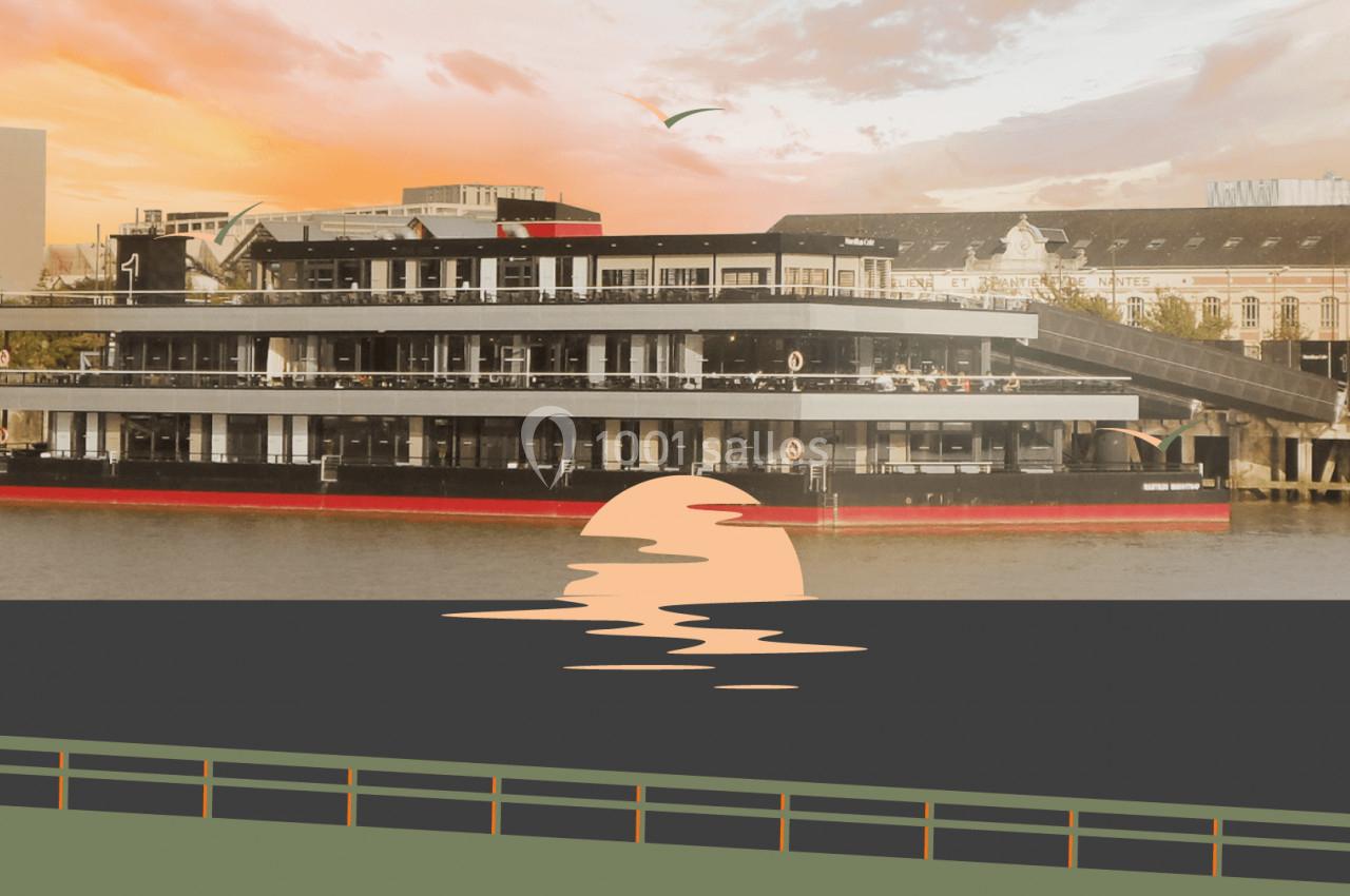 Bateau moderne à plusieurs étages amarré sur une rivière au coucher du soleil, avec un bâtiment en arrière-plan.
