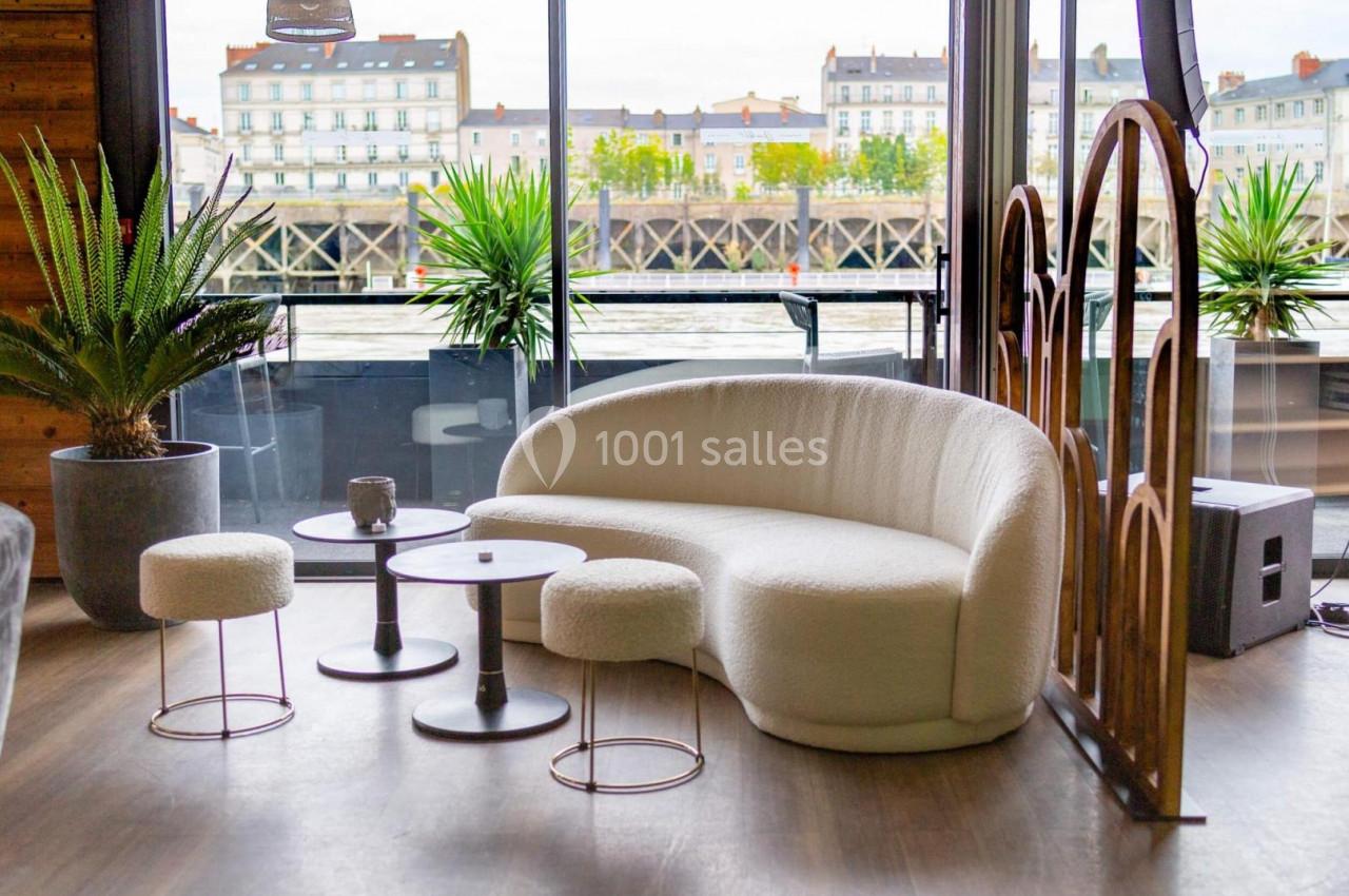 Salon moderne avec canapé blanc incurvé, tabourets assortis, tables basses et vue sur des bâtiments au bord de l'eau.