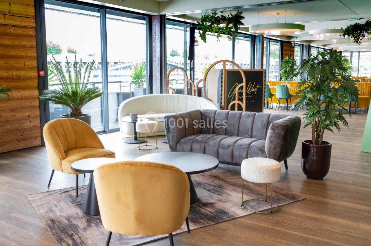 Espace intérieur lumineux avec mobilier moderne, fauteuils jaunes, canapé gris et plantes décoratives près de grandes baies…