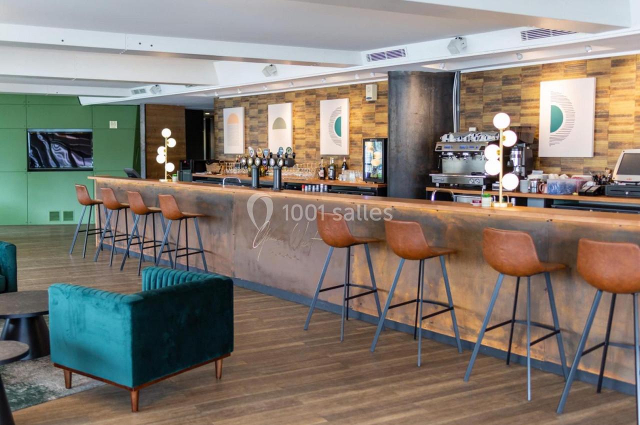 Bar moderne avec comptoir en métal, tabourets en cuir, fauteuils verts et décorations murales géométriques.
