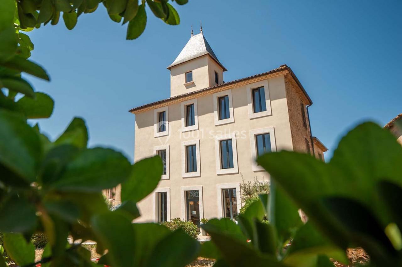 Façade d'un bâtiment ancien rénové avec une tour centrale, vue à travers des feuillages sous un ciel bleu.
