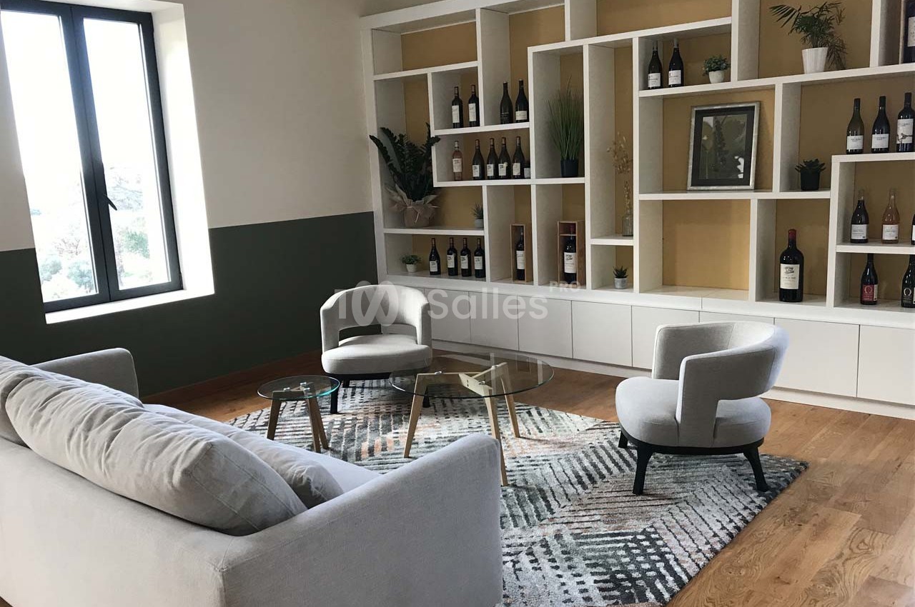 Salon moderne avec canapé, fauteuils, table basse en verre, étagères décorées de bouteilles et plantes, parquet au sol.