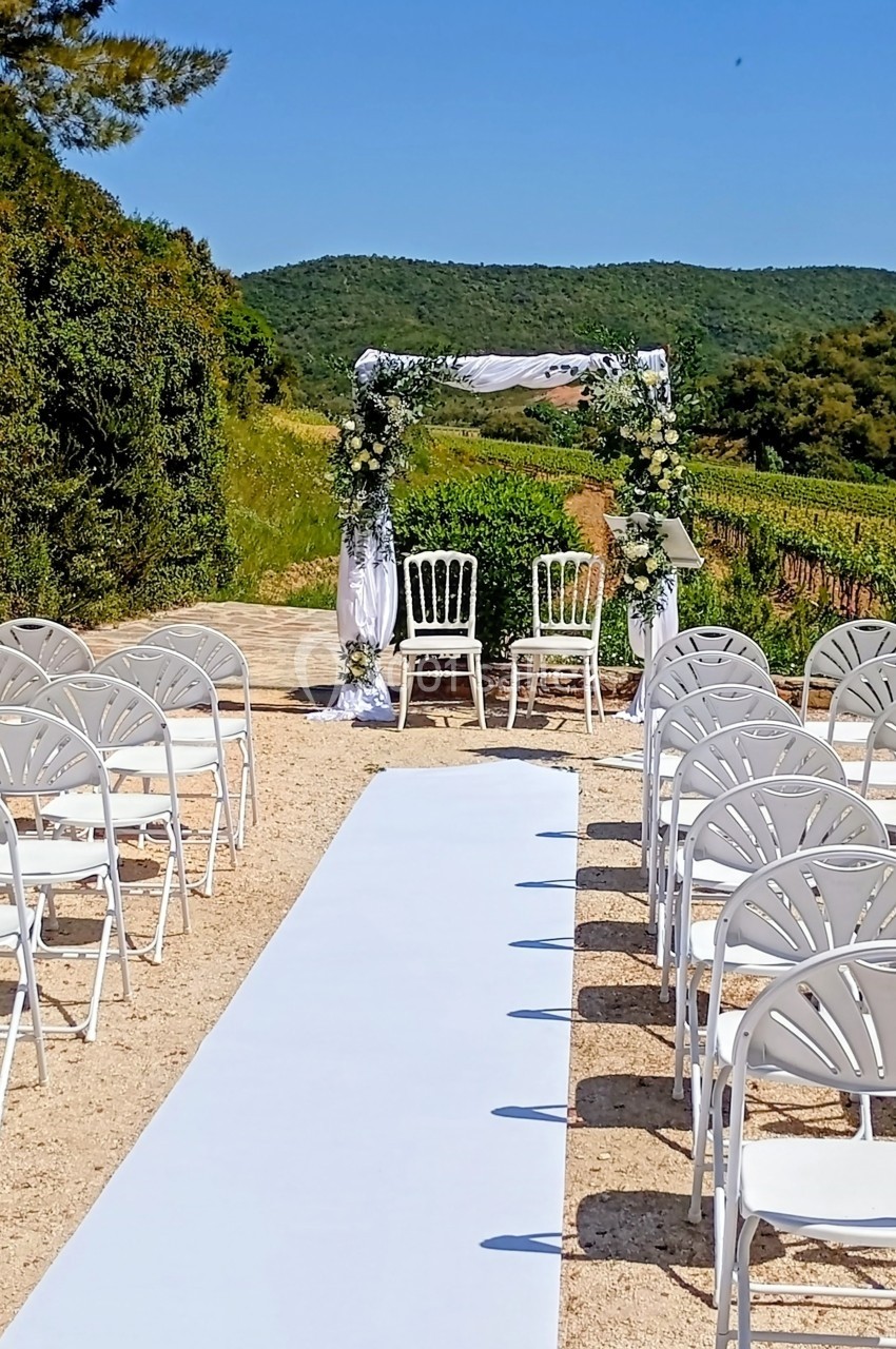 Allée centrale blanche menant à une arche fleurie avec deux chaises, entourée de rangées de chaises blanches en extérieur.