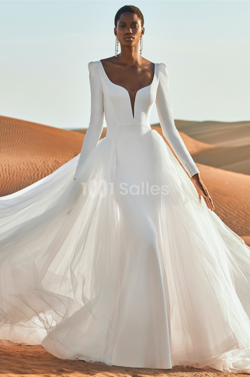 Femme portant une robe de mariée blanche fluide avec manches longues, posant dans un désert sous un ciel clair.