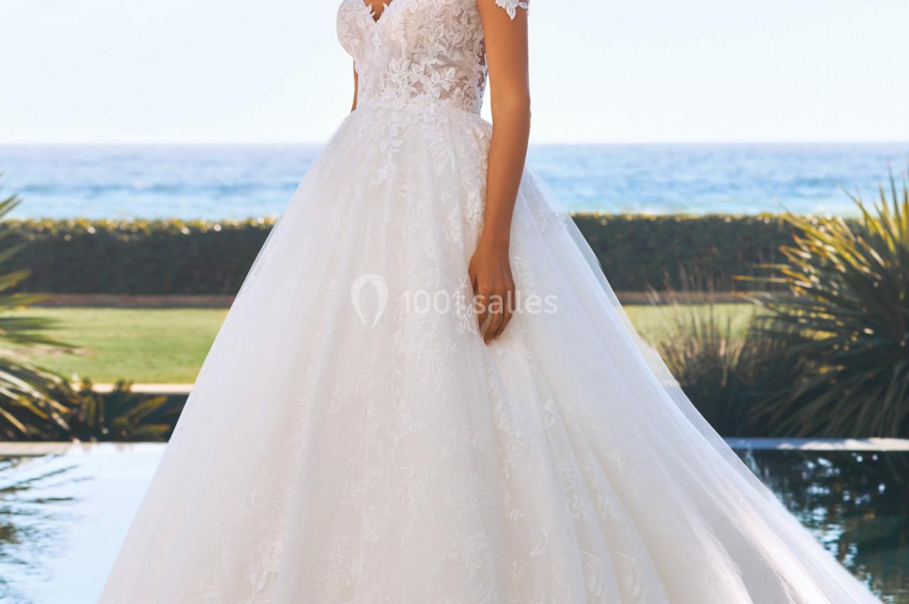 Femme portant une robe de mariée blanche avec dentelle, posant en extérieur près d'une piscine et d'un paysage côtier.