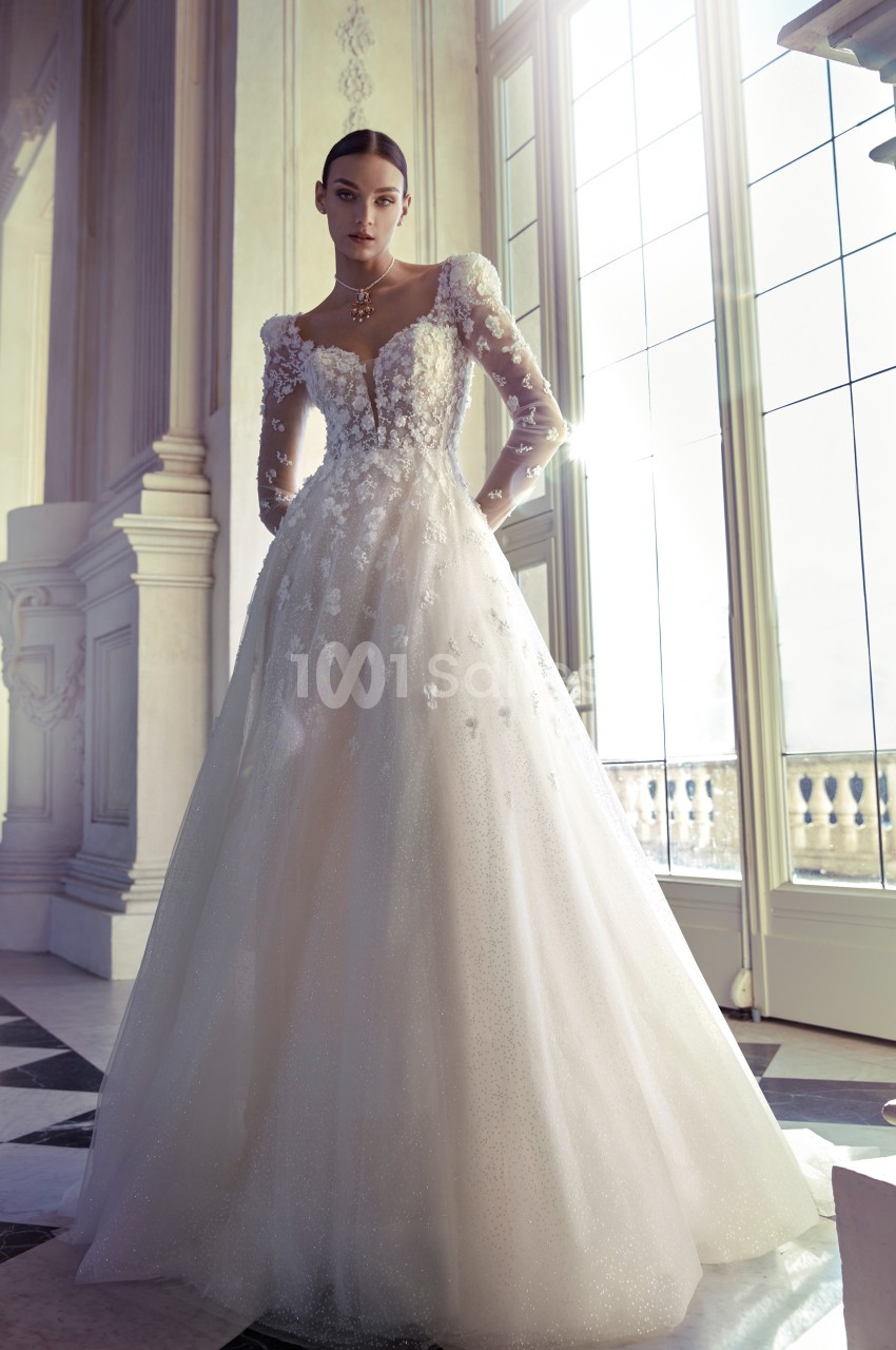 Femme portant une robe de mariée élégante avec manches longues en dentelle, dans une pièce lumineuse avec de grandes…