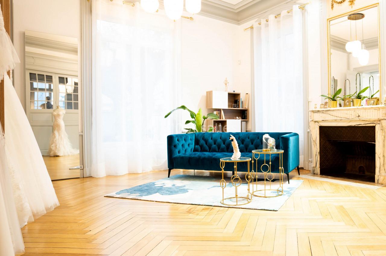 Salon lumineux avec parquet en chevrons, canapé bleu, tables dorées, miroir doré et plantes décoratives.