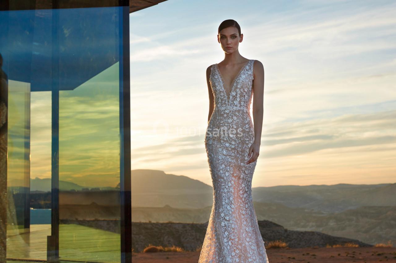 Femme portant une robe de mariée ornée de dentelle, debout en extérieur au coucher du soleil près d'une structure moderne.