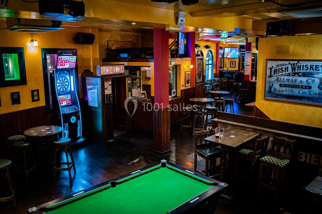 Salle de bar avec billard, cible de fléchettes, tables et chaises en bois dans une ambiance chaleureuse et tamisée.